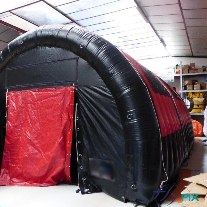 Une tente gonflable en forme de tunnel, en enveloppe PVC 0.6 mm et double peau capitonnée, de couleur noire avec des portes rouges.