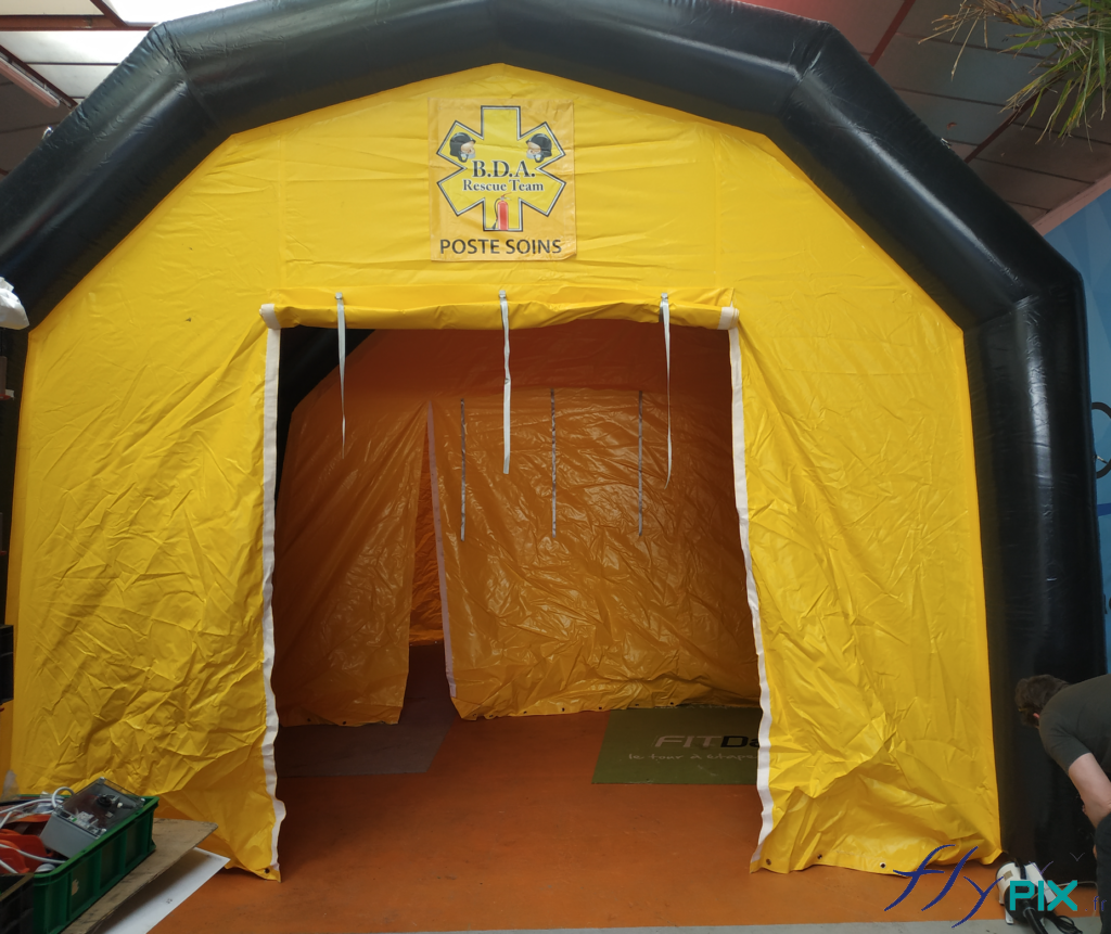 Une vue de face de la tente PMA: porte large pignon, avec fixations velcro, banderole de marquage imprimée, fixations velcro au-dessus de la porte, boudins d’ossatures air captif de couleur noir, gonflé à l’air avec une pompe électrique.
