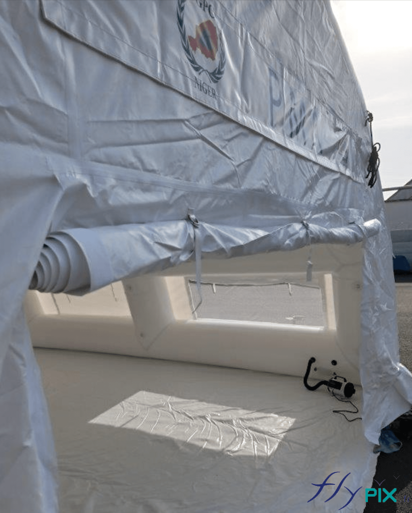 Une vue détaillée de la porte large pignon de la tente médicale gonflable air captif. On voit aussi la bâche au sol en PVC lavable. Sur cette photo, nous pouvons observer: la large porte pignon ouverte, la banderole de marquage amovible imprimée fixée par velcro, des larges fenêtres (avec moustiquaires et volets), et en bas à droite la pompe régulateur de pression. Le régulateur de pression assure que la pression est toujours identique à l’intérieur des boudins d’ossatures, qui sont aussi dotés de valves de surpression.