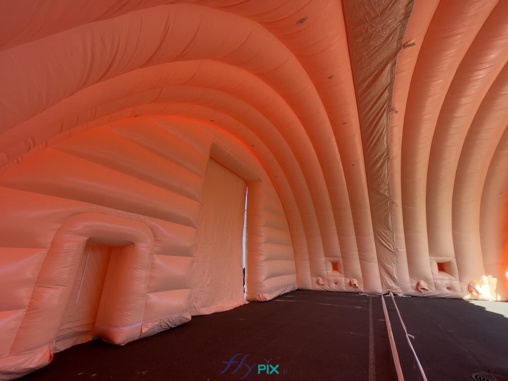 Une prise de vue photo générale qui montre 2 modules de hangar gonflable militaire assemblés et connectés, avec le raccordement par l'intérieur qui assure l'étanchéité de la toiture en cas de pluies ou d'intempéries. À gauche, nous voyons la petite porte secondaire de service, ensuite la grande porte d'accès principale, sur le pignon avant. Au fond, sur chaque module, dans la partie inférieure, nous pouvons observer les fenêtres et les manchons à air et les trous passe fils et gaines électriques. - Copyright : Vivien LAÏLLE/FLYPIX, droits réservés. Une prise de vue photo générale qui montre 2 modules de hangar gonflable militaire assemblés et connectés, avec le raccordement par l'intérieur qui assure l'étanchéité de la toiture en cas de pluies ou d'intempéries. À gauche, nous voyons la petite porte secondaire de service, ensuite la grande porte d'accès principale, sur le pignon avant. Au fond, sur chaque module, dans la partie inférieure, nous pouvons observer les fenêtres et les manchons à air et les trous passe fils et gaines électriques. - Copyright : Vivien LAÏLLE/FLYPIX, droits réservés.